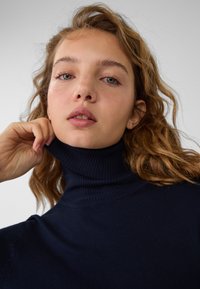 Navyblauer Stehkragenpullover aus geripptem Strickstoff, der einen hohen Kragen und eine figurbetonte Passform aufweist. Das Modell passt den Kragen mit einer Hand an.