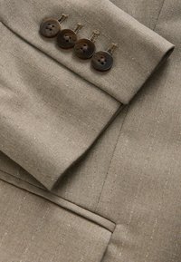 Beige tailored jacket sleeve featuring four dark buttons, subtle pinstripes, and textured fabric with visible stitching detail.