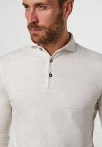 Hombre barudo que lleva una camiseta polo de manga larga color beige claro con dos botones oscuros abrochados, mirando hacia un lado contra un fondo sencillo.