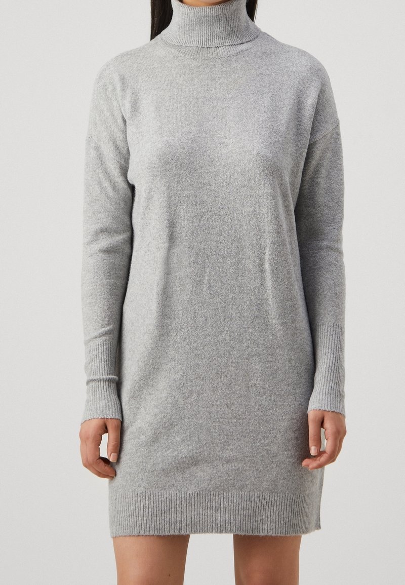 Femme portant une robe-pull à col roulé côtelée gris clair, manches longues, coupe droite, visible du cou jusqu'à mi-cuisse.