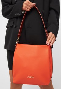 Oranje leren handtas met een gestructureerde afwerking, voorzien van een bruine verstelbare schouderband en gouden hardwaredetails, inclusief het logo aan de onderkant.