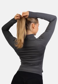 Dopasowana bluzka z długim rękawem w ciemnoszarym kolorze, z wysokim kołnierzem i otworami na kciuki w mankietach, noszona podczas związania włosów w kucyk.