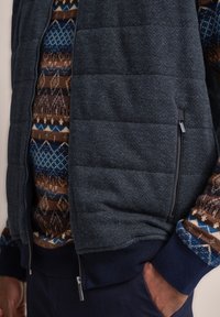 Grauer Fischgrät-Puffer-Weste mit einer Reißverschlusstasche, geschichtet über einem gemusterten Strickpullover in Blau- und Brauntönen. Rippbündchen und -saum.