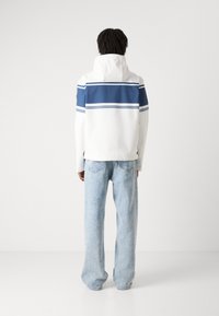 Hvid jakke med blå vandrette striber, med hætte og afslappet pasform. Bæres med lyseblå brede jeans. Minimalistisk design.
