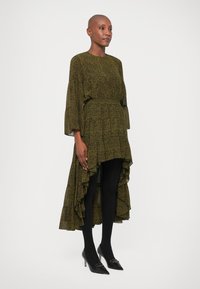 Robe verte à motifs avec des manches ballon, ourlet asymétrique et accents ondulés ; associée à des collants noirs et des escarpins à bout pointu.