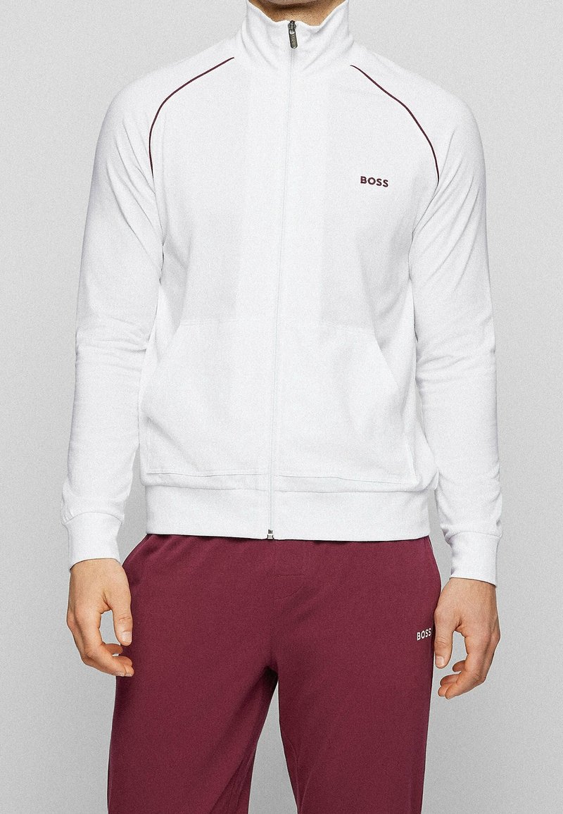 Giacca bianca con cerniera e colletto alto, dettagli bordeaux e logo. Presenta maniche lunghe e una texture morbida, abbinata a pantaloni color bordeaux.