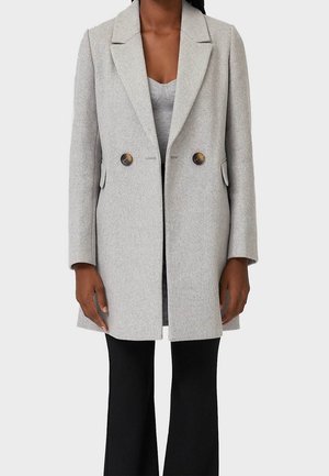 Woman wearing a mid-length light gray double-breasted coat with dark buttons, a gray top, and black pants against a plain background.