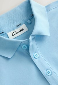 Hellblaues Polohemd mit drei Knöpfen und umgeschlagenem Kragen, mit einem weißen Etikett mit dem Schriftzug "Clarks" im Halsausschnitt.