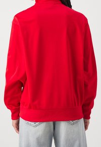 Czerwona sportowa bluza z wysokim kołnierzem, z długimi rękawami i prążkowanym ści ągaczem. Tył ma gładką fakturę, bez widocznych wzorów czy logo.