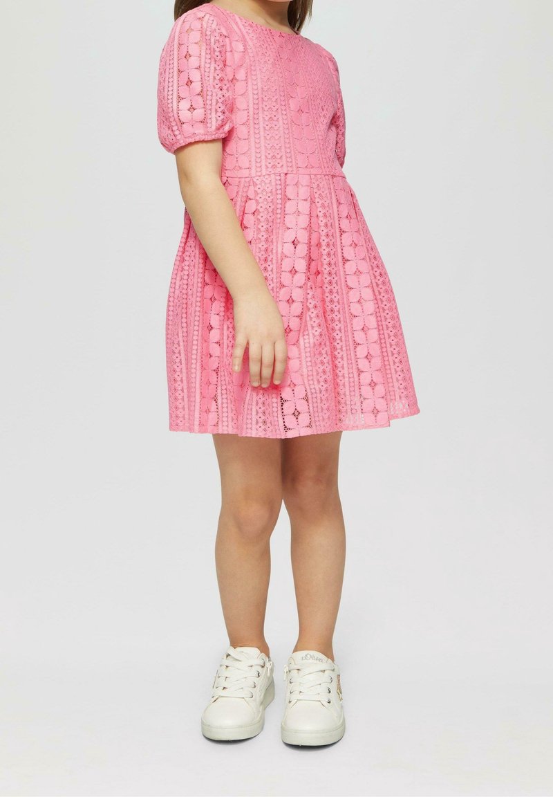 Robe rose avec des manches courtes bouffantes, présentant des motifs en dentelle et une jupe évasée. Portée avec des baskets blanches, mettant en avant un look décontracté.