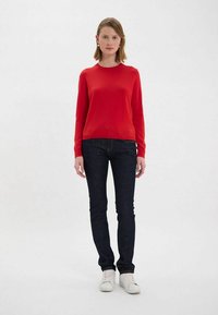 Pull en tricot rouge à manches longues avec col rond, associé à un jean ajusté bleu foncé et des baskets blanches. Texture lisse, design minimaliste.