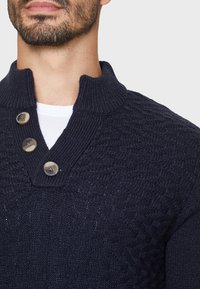 Marineblau gestrickter Pullover mit einem strukturierten Muster, der über einen gerippten Schalkragen und drei Knöpfe an der Leiste verfügt. Material aus Baumwollmischung.