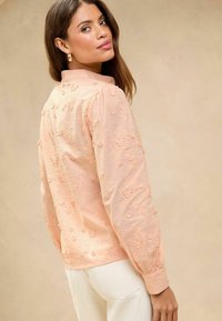 Blusa imbottita color pesca chiaro con ricami a fiori in 3D, colletto con bottoni e maniche lunghe, abbinata a pantaloni color crema.