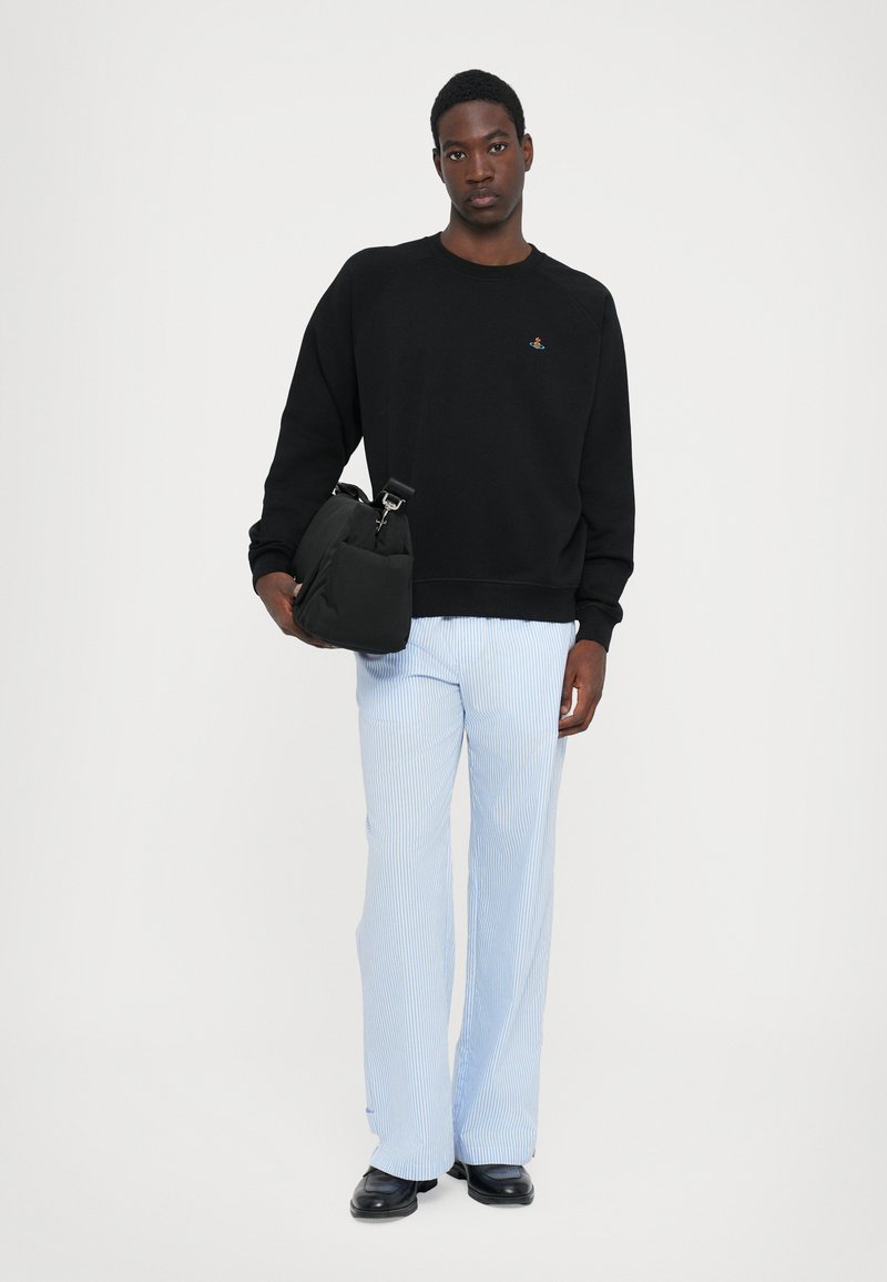 Schwarzes Sweatshirt mit Rundhalsausschnitt und buntem Logo, kombiniert mit hellblauen gestreiften weitem Hosen. Schwarze Tasche mit verstellbarem Tragegurt.