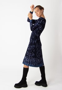 Robe marine avec un motif floral léger bleu complexe, manches longues, taille cintrée, jupe plissée, associée à des chaussettes noires jusqu'aux genoux et des bottes noires chunky.