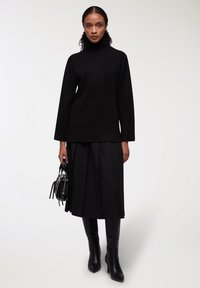 Czarny sweter z golfem i długimi rękawami, zestawiony z czarną plisowaną spódnicą i czarnymi kozakami za kolano. Nosi małą czarną torebkę.