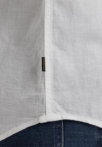 Camisa de lino blanca con dobladillo curvado, que presenta una etiqueta en la costura lateral. La tela parece ligera y ligeramente texturizada, en contraste con el denim oscuro.