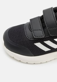 Czarne buty sportowe z siateczkową cholewką, wyłożonym językiem i dwoma paskami na rzepy. Białe akcenty po bokach. Teksturowana gumowa podeszwa.