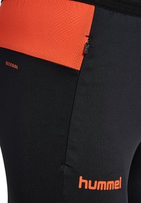 Czarne legginsy sportowe z teksturowanym, pomarańczowym paskiem w talii, wyposażone w małą kieszonkę na zamek oraz pomarańczowe logo "hummel" po boku.