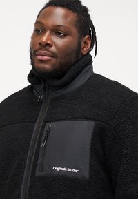 Μαύρο φλις μπουφάν με ψηλό γιακά, κλείσιμο με φερμουάρ και τσέπη στο αριστερό στήθος που διαθέτει μαύρο πάνελ υφάσματος και λεπτομέρεια λογότυπου.