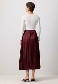 Jupe plissée bordeaux longue en tissu fluide, associée à un haut blanc ajusté à manches longues. Des sandales à talons noires complètent le look.