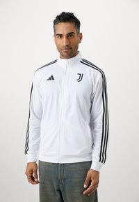 Hvid zip-up jakke med sorte striber på ærmerne, Adidas-logo og Juventus-emblem. Fremstillet af glat, letvægtsmateriale.