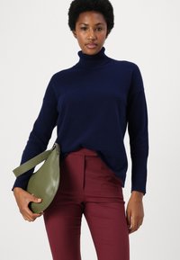 Suéter de cuello alto en color navy de material suave, combinado con pantalones ajustados burdeos. Sosteniendo un bolso circular de cuero verde con una correa.