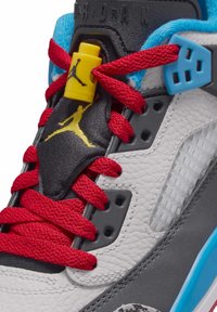 Dettaglio di una sneakers Air Jordan bianca e grigia con lacci rossi, logo Jumpman giallo sulla linguetta nera e fermaglio per lacci blu.
