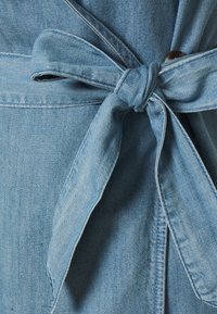 Světle modrý denimový kousek oděvu s uvázaným pasem na uzel, vykazující hladkou texturu a prošívané akcenty podél okrajů.