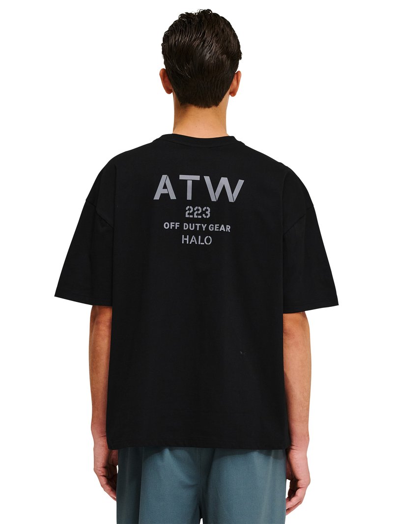 Mężczyzna noszący czarny, oversize'owy T-shirt z napisem „ATW 223 OFF DUTY GEAR HALO” na plecach, w połączeniu z niebieskimi spodniami w kratę.