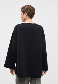 Volně střižený svetr s dlouhým rukávem z tmavě námořnické pleteniny. Má široký výstřih a zaoblený lem. Nošený přes šedé kárované kalhoty.