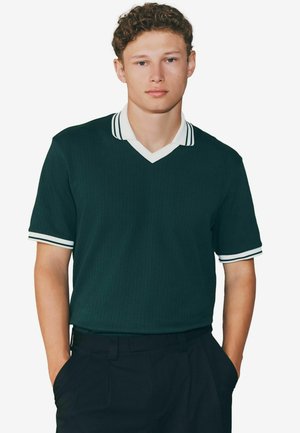 Camiseta polo de manga corta color verde oscuro con escote en V, que presenta un collar blanco y acentos a rayas en las mangas. Tejido texturizado con un acabado acanalado.
