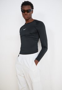 Cămașă sport lungă, neagră, cu un fit ajustat și logo-ul alb Nike, asortată cu pantaloni cargo gri deschis, prevăzuți cu buzunare laterale.