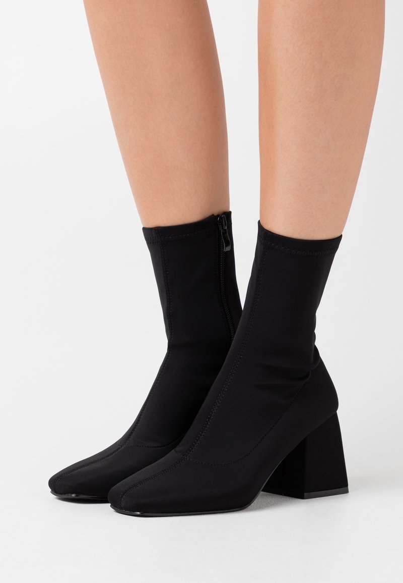 Botas de tobillo negras hechas de tela elástica, con punta afilada y tacón cuadrado. Cierre lateral con cremallera y detalles de costura visibles.