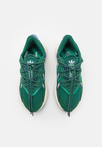 Par av gröna Adidas-sneakers med lila och teal accenter, vita sulor och fastsatta med mönstrade snören på en vit bakgrund.