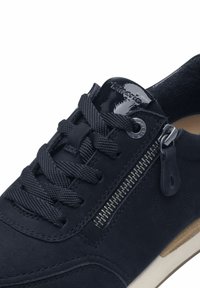 Sneaker in suede nera con punta rotonda, lacci testurizzati, dettaglio con zip laterale e suola beige a contrasto. Gli accessori includono una linguetta lucida.