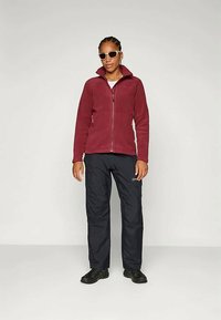 Casaco de fleece bordeaux com fecho de zíper integral, combinado com calças pretas isoladas. O modelo usa óculos de sol brancos e sapatos pretos. Design simples.
