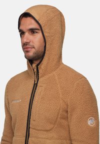 Felpa con cappuccio in pile color tan, con esterno testurizzato, chiusura a zip frontale e rifiniture nere a contrasto. Presenta un cappuccio arrotondato e un dettaglio del logo sulla manica sinistra.