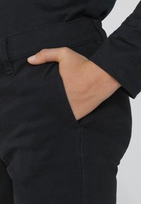 Černé bavlněné kalhoty s hladkým povrchem, se spodní kapsou a přiléhavým pasem. Ruka leží v kapse.