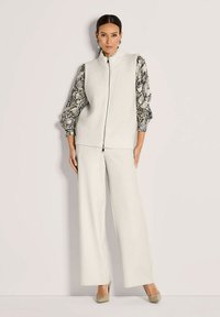 Frau in einem weißen, sowie aufreißbarem Strickwesten über einer Bluse mit Schlangenmuster, breiten weißen Hosen und beigen spitzen Absätzen, steht vor einem schlichten Hintergrund.