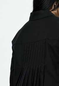 Czarna koszula z plisowanym wzorem na plecach. Gładka tkanina z usztywnianym kołnierzem i czystymi liniami.