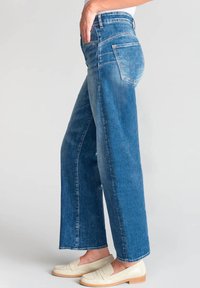 Jean large en denim bleu moyen avec une finition délavée, présentant une taille mi-haute, cinq poches et une coupe droite.