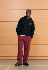 Fekete kapucnis pulóver rózsaszín geometriai logóval, bordó melegítőnadrággal és fekete sportcipővel párosítva. A háttérben fa panelek láthatók.