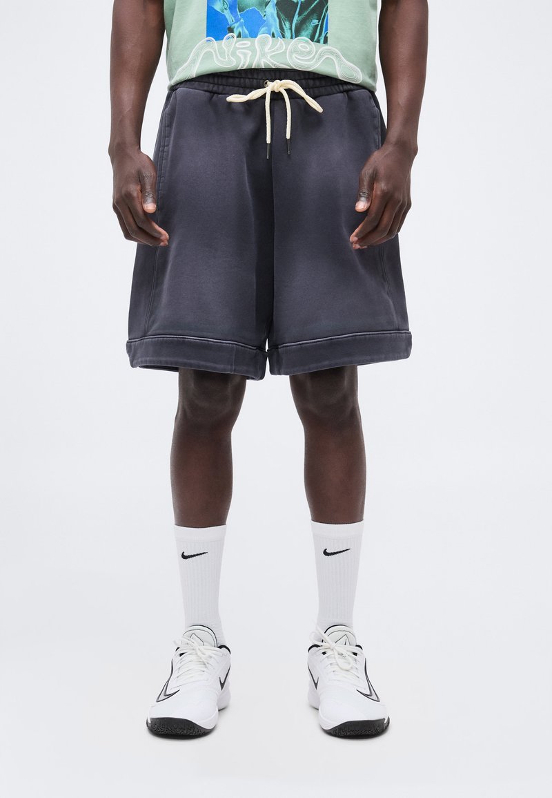 Man die zwarte short met trekkoord, witte Nike sokken en witte Nike sneakers draagt tegen een effen lichte achtergrond.