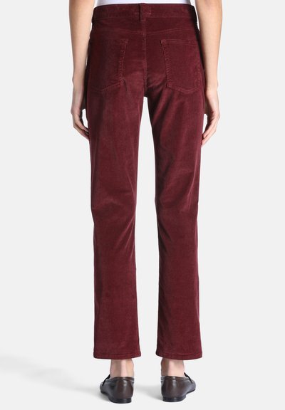 Personne portant un pantalon droit en velours côtelé rouge foncé et des mocassins marron foncé, vue de dos sur un fond uni.
