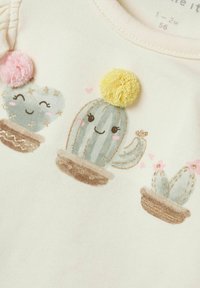 Biała tkanina z kreskówkowymi kaktusami z teksturowanymi pomponami w kolorze różowym i żółtym. Złote akcenty i uśmiechy przedstawione na kaktusach.