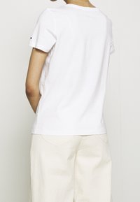 Camiseta blanca de manga corta con cuello redondo, hecha de algodón suave. Presenta un acento de logotipo en la manga izquierda, combinada con pantalones de colores claros.