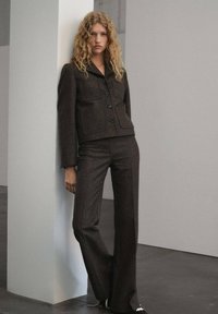 Mujer con cabello rubio rizado, vistiendo una chaqueta de botones marrón oscuro y unos pantalones anchos a juego, apoyada contra un pilar blanco en interiores.