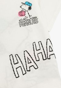Camiseta blanca de algodón con un dibujo de un perro de cartoon sosteniendo un objeto rosa, con el logo de "PEANUTS" y el texto negro "HAHA" en una fuente ondulada.