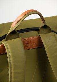 Groene stoffen tas met leren accenten, voorzien van een bovenhandvat, verstelbare schouderbanden en een logo-patch gestikt op een beige leren gedeelte.